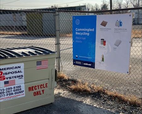 Recycling Sign hanging on a fence next to a front loading waste bin